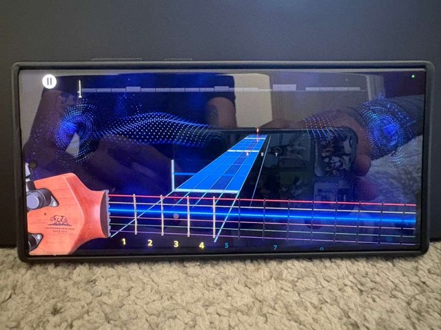 Phone in landscape showing Rocksmith+ gameplay with scrolling notes on the virtual fretboard